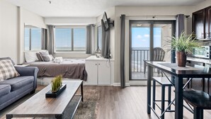 A modern apartment with a living room, dining area, and kitchenette. It features a large window with blinds, a sofa, a coffee table, and a TV mounted on the wall.