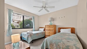 Un dormitorio con dos camas, un ventilador de techo, un aparador, una lámpara y una ventana con cortinas.