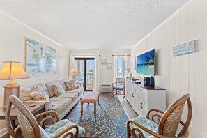 A room with a sofa, chairs, a television, and artwork on the walls. - Ocean View Escape | Steps to Ocean | Relax Now | OFV E206 (Myrtle Beach)