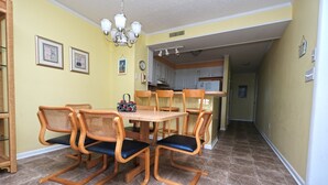 A dining area with a round table and six chairs, a chandelier, and a kitchen in the background. - Pelican Landing 125 - Myrtle Beach (Myrtle Beach)