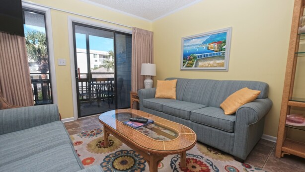 A living room with a sofa, a glass-top coffee table, a patterned rug, a painting on the wall, and a sliding glass door leading to an outdoor area. - Pelican Landing 125 - Myrtle Beach (Myrtle Beach)