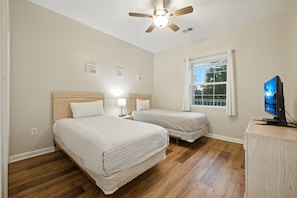 Quarto com duas camas, um ventilador de teto, uma janela com cortinas e uma televisão em um armário.