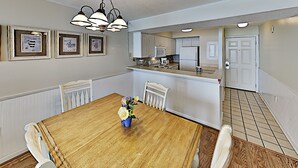 Ein Esszimmer mit Holztisch, weißen Stühlen und einem Blumenstrauß. An der Wand hängen gerahmte Bilder, im Hintergrund ist eine Küche zu sehen und rechts eine Tür.