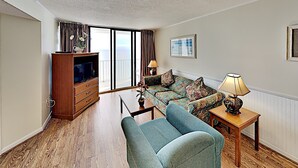 Ein Wohnzimmer mit einer Couch, einem Fernseher, einer Lampe und Meerblick durch große Fenster.