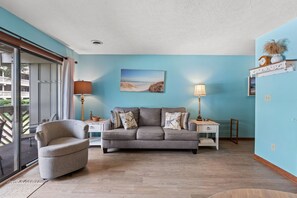 A living room with a grey sofa, a grey armchair, a wooden coffee table, two floor lamps, a painting of a beach, and a view through a glass door.