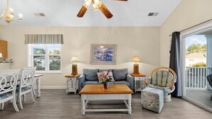 A spacious living room with a wooden coffee table, a sofa, a dining table with chairs, and a ceiling fan. - Willowbend 831 - Barefoot (North Myrtle Beach)