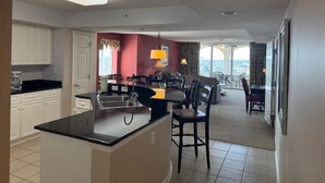 A modern kitchen with white cabinets, a black countertop, and a central island. There is a dining area with a table and chairs, and a view through a glass door.