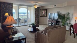 A hotel room with a sofa, a TV, a dining table, a lamp, a plant, and a balcony with a view.