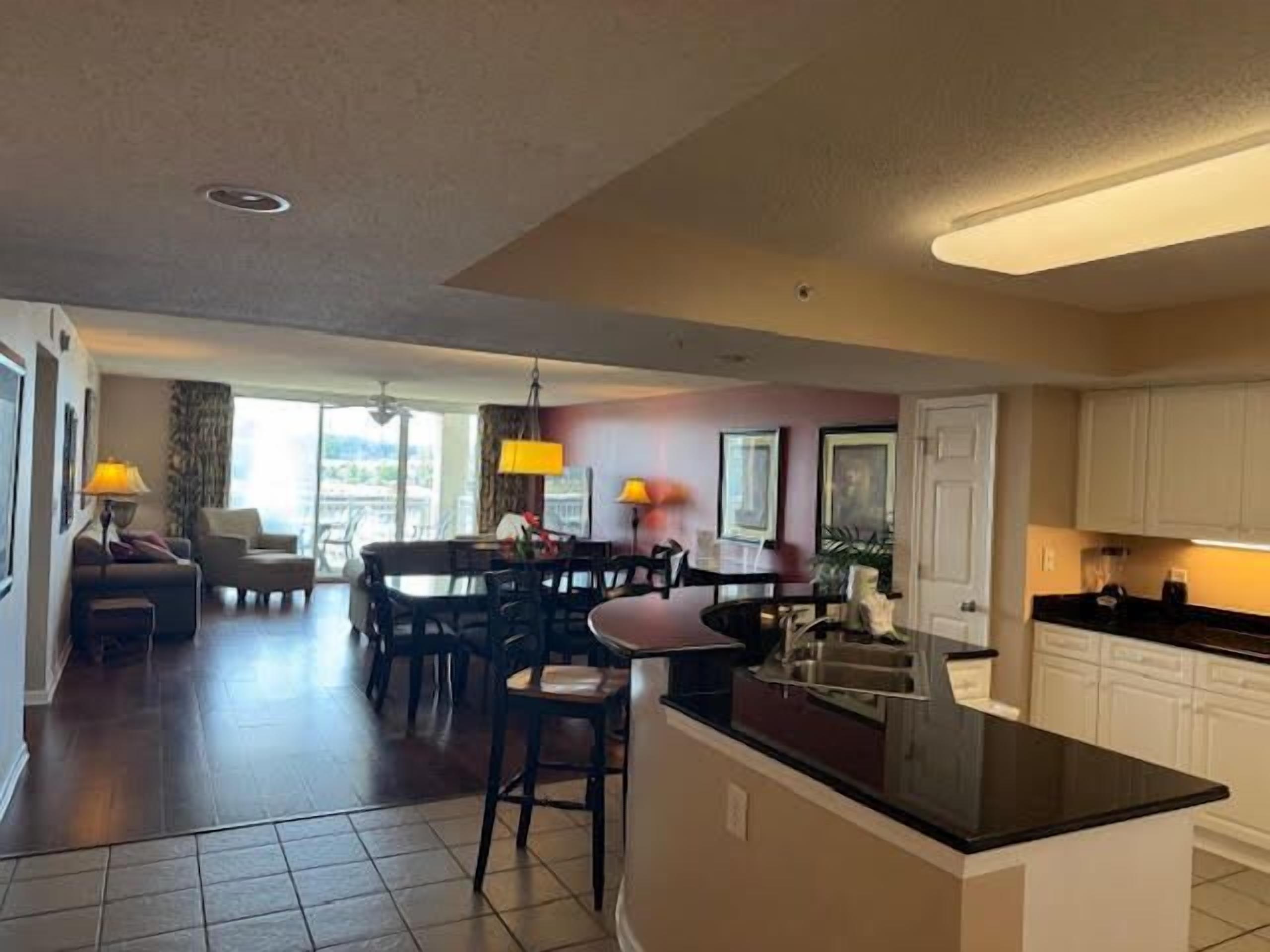 A modern kitchen with a central island, dining area with chairs, and a living room with a sofa and lamp.
