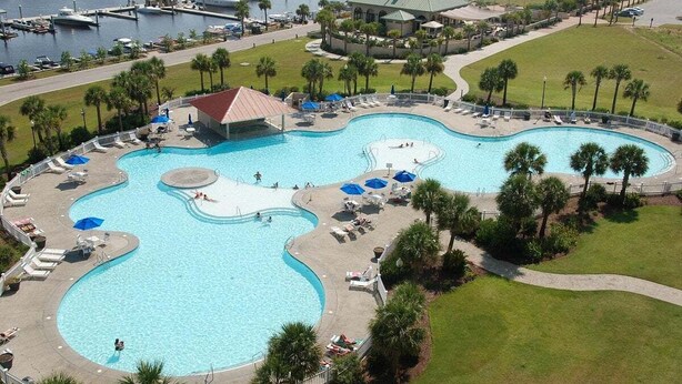 A large, multi-pond pool area with lounge chairs and umbrellas, surrounded by palm trees and a marina in the background.