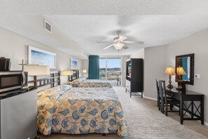A hotel room with a bed, a desk, a chair, a TV, a microwave, a ceiling fan, and a view of the outside.
