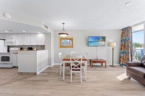 A modern kitchen with white cabinets, a dining area with a wooden table and chairs, a flat-screen TV mounted on the wall, and a view of the ocean through large windows.