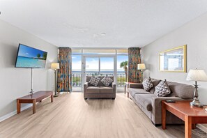 A living room with a sofa, a television, a bench, and a view of the ocean through large windows.