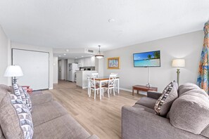 A modern living room with a sofa, a dining table with chairs, a kitchen area, and a TV mounted on the wall. - Atlantica 154, Beachfront Myrtle Beach Sping Retreat (Myrtle Beach)