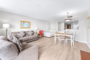 A modern living room with a sofa, a dining table with chairs, a bed, and a kitchen area in the background.