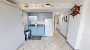 A compact kitchen with blue cabinets, a white refrigerator, and a microwave. There are decorative wall stickers of a crab, a shark, and a ship's wheel. A trash bin is placed near the entrance. - Atlantica 301, Oceanfront Myrtle Beach Condo Rental (Myrtle Beach)