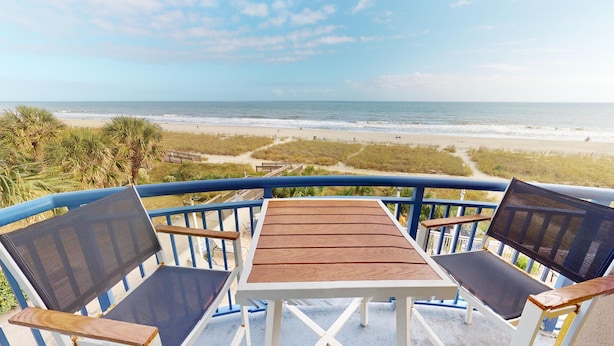 A balcony with a wooden table and two chairs, overlooking a beach with a fence and palm trees. - Atlantica 301, Oceanfront Myrtle Beach Condo Rental (Myrtle Beach)