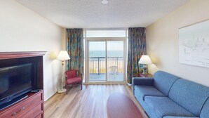 Habitación de hotel con televisión, sofá, silla, dos lámparas y balcón con vista a la playa.