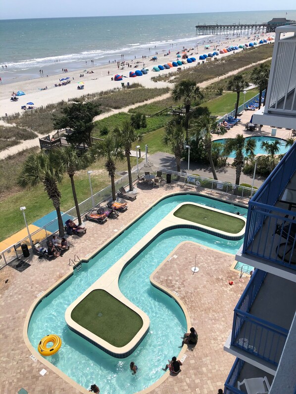Uma área de piscina com várias piscinas, uma praia com guarda-sóis e um píer ao fundo.