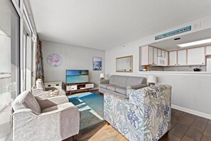 Sala de estar com sofá, poltronas, televisão, mesa de centro e área de cozinha com armários e micro-ondas.