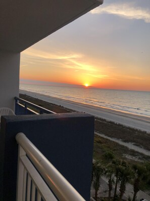 Uma varanda com vista para o pôr do sol sobre o oceano, uma praia de areia e palmeiras.