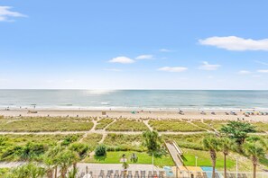 A beach view with a clear sky, a sandy beach, and a pool area with lounge chairs.