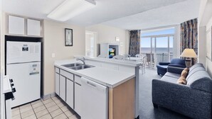 A compact kitchen with a sink, a refrigerator, and a dishwasher. There is a dining area with a blue couch and a table. - Atlantica 1202, Myrtle Beach Ocean Escape (Myrtle Beach)
