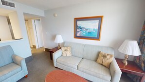 A hotel room with a light blue sofa, a wooden coffee table, a painting of a coastal scene, and a lamp on a side table.