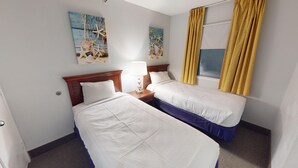 A hotel room with two single beds, a wooden headboard, a nightstand with a lamp, and two wall art pieces.