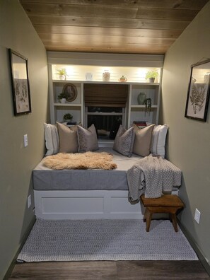 Une chambre confortable avec un lit, des oreillers, un petit tabouret et une fenêtre avec des stores.