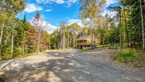 A cabin with a gravel driveway leading to a wooden deck, surrounded by tall trees and a clear sky. - Red Fox Lodge - Close to downtown Rangeley w/ Hot Tub & Sauna (Rangeley)