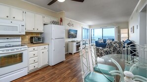 A compact kitchen with white appliances, a dining area with a glass table and teal chairs, and a view of the ocean through large windows. - Oceanfront 2BD- Cherry Grove Point- Ocean View K3 (North Myrtle Beach)