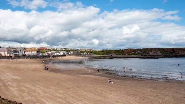 En strand med en sandstrand, några byggnader i bakgrunden och en klar himmel med moln.