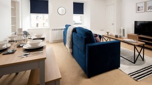 Ett modernt vardagsrum med en blå soffa, ett matbord dukat med vita tallrikar, glas och bestick. Det finns en tv, ett element och ett fönster med en mörk persienn.
