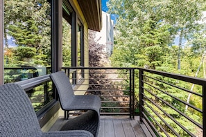 Un balcon avec des fauteuils en osier, une terrasse en bois et une vue sur les arbres et un immeuble.