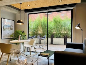 A modern interior with a dining area, a sofa, and large windows with a view of greenery and a brick building.