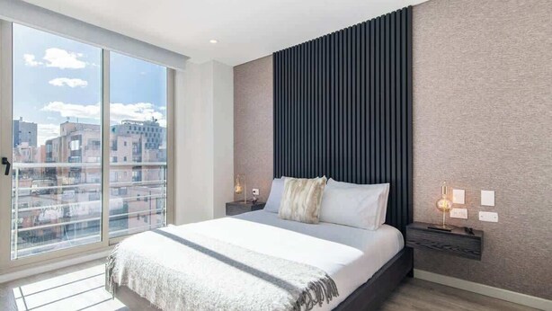 Ein modernes Schlafzimmer mit einem großen Bett, Blick auf die Stadt durch raumhohe Fenster und einem dunklen Kopfteil.