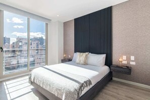 Un dormitorio moderno con una cama grande, vistas a la ciudad a través de ventanas de techo a piso, y un cabecero oscuro.