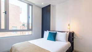 Ein modernes Hotelzimmer mit großem Fenster, einem Bett mit weißen Leinentextilien und einem türkisfarbenen Kissen, einem dunklen Kopfteil und einer Nachttischlampe.