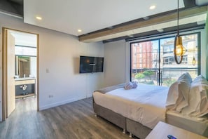 A modern bedroom with a large bed, a TV mounted on the wall, and a window with a view of buildings.