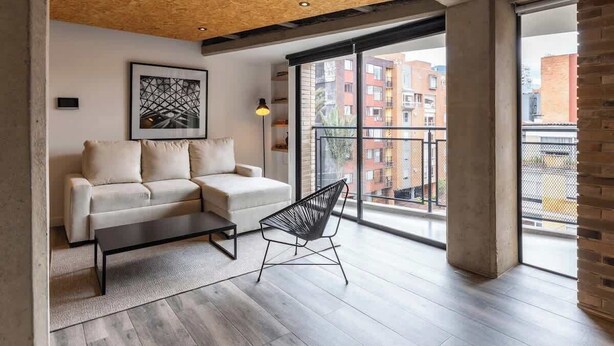 Ein modernes Wohnzimmer mit einer Couch, einem Sessel, einem gerahmten Kunstwerk und einem großen Fenster mit Blick auf Gebäude.