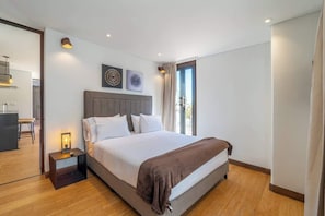 Un dormitorio moderno con una cama grande, mesitas de noche, una mesilla y un cabecero, junto a una ventana con cortinas.