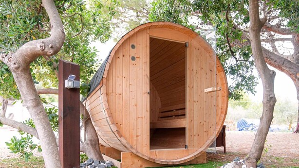 Uma estrutura em forma de barril de madeira, com uma porta aberta, cercada por árvores e grama.