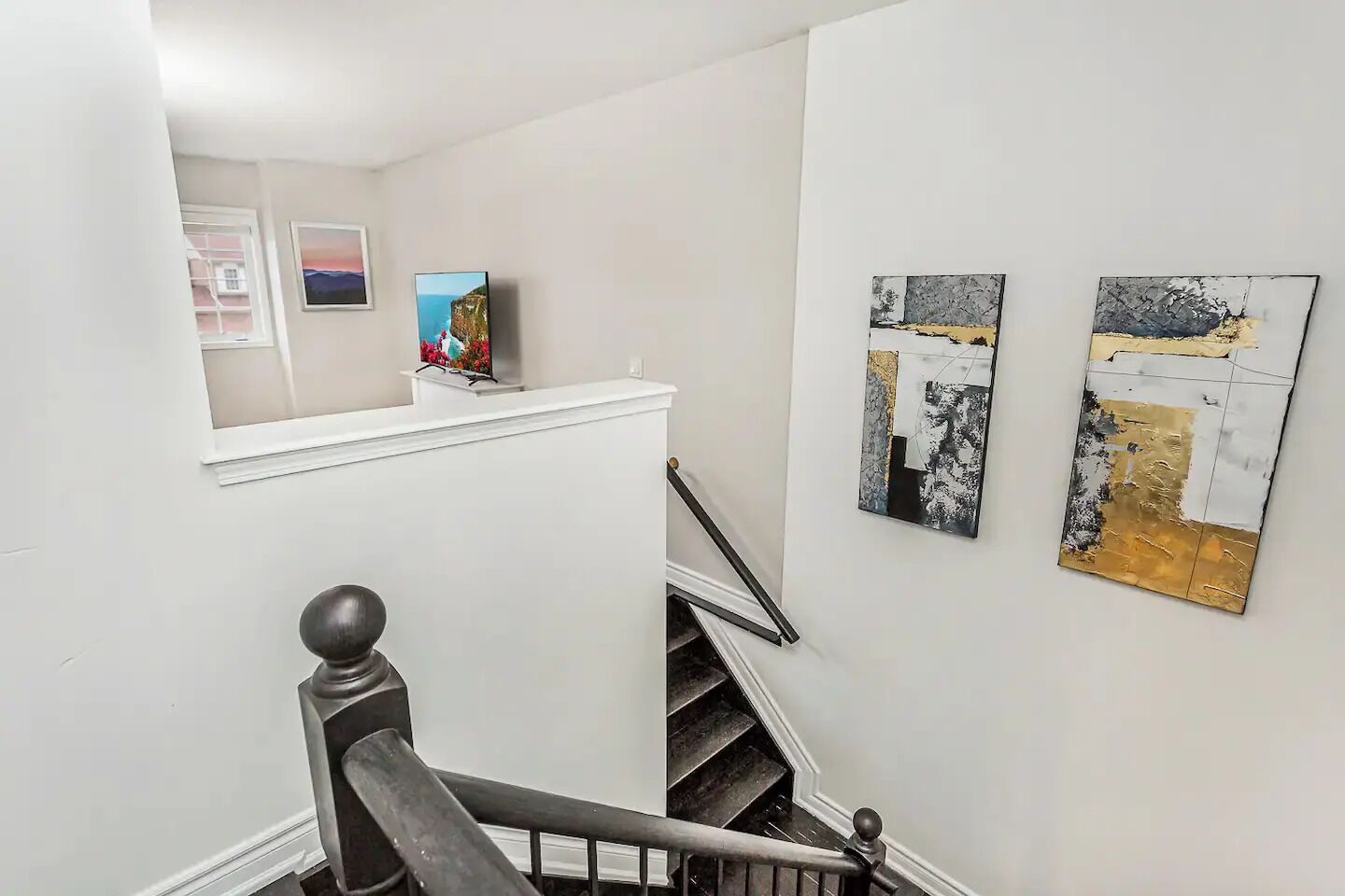 Una escalera con barandilla, un pequeño estante con un cuadro enmarco, y dos pinturas abstractas en la pared.