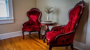 Two red velvet armchairs with wooden frames, a small round table with a white vase, and a wooden floor in a room with a window and beige walls. - Tranquil TN Escape! Book Your Stay In Elizabethton (Elizabethton)