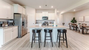 A modern kitchen with white cabinets, stainless steel appliances, a central island with stools, and a dining area with a wooden table and chairs. - 5BR WindMark Beach Bunk Room, Pools, Golf Cart, Ping Pong  (Port St. Joe)
