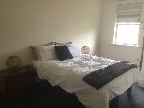 A neatly made bed with white linens and a dark throw blanket, a bedside lamp, and a wicker basket on a nightstand. - Quite cosy Cottage close to Port. Pet friendly (Port Albert)