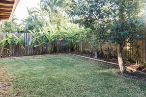 Ein Garten mit Holzzaun, Rasen und verschiedenen Pflanzen, darunter Bananenstauden und eine Palme.
