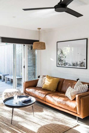 A modern living room with a leather sofa, a round coffee table, a ceiling fan, and a large window. - The Sunshine Bungalow Beautiful Noosa Home (Noosa)