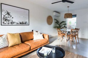 A living room with a brown leather sofa, a round coffee table, and a dining area with wooden chairs and a table.